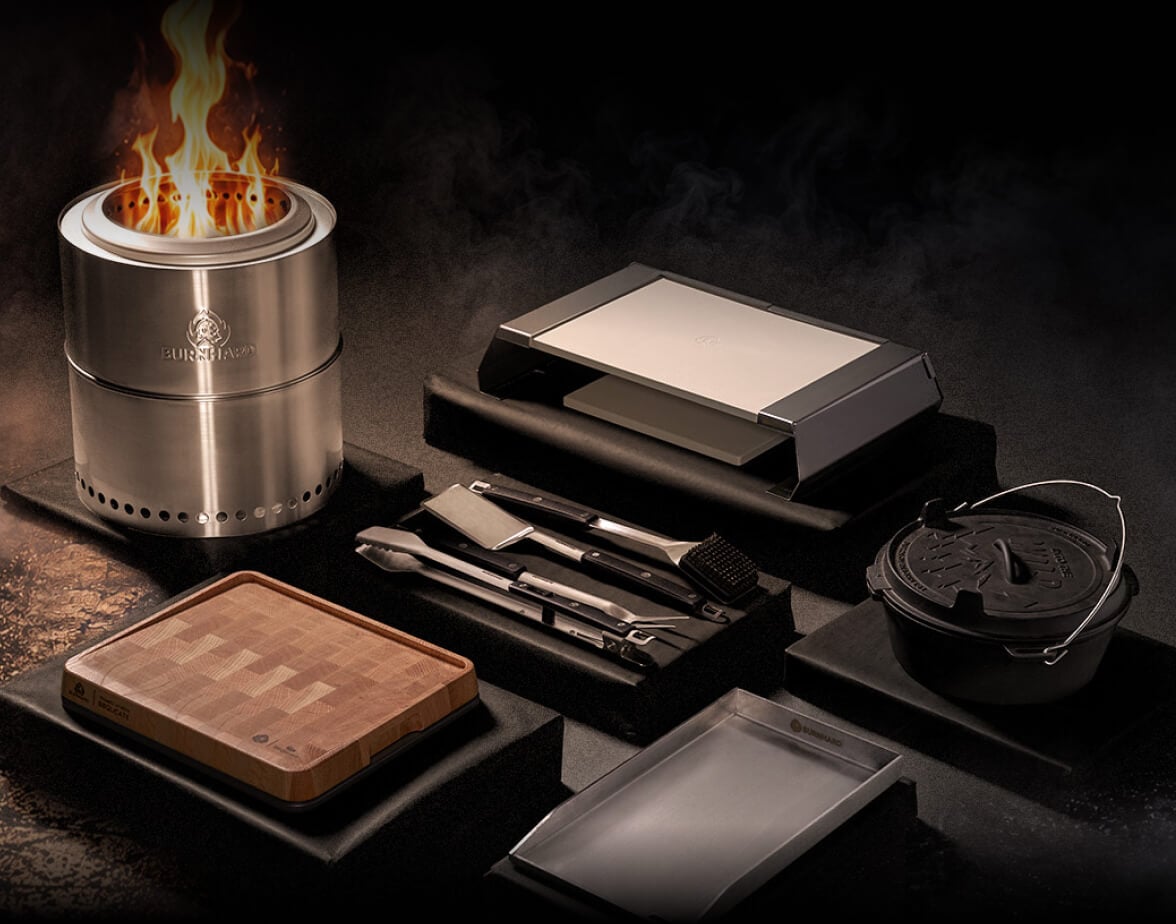 Feuerkorb mit brennendem Feuer, Grillzubehör, Kontaktgrill, Dutch Oven und Metallschale auf dunklem Untergrund.